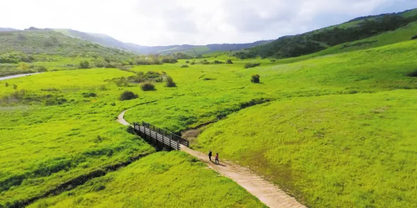 A dirt path winds through bright green hills to a small wooden bridge with two people walking.