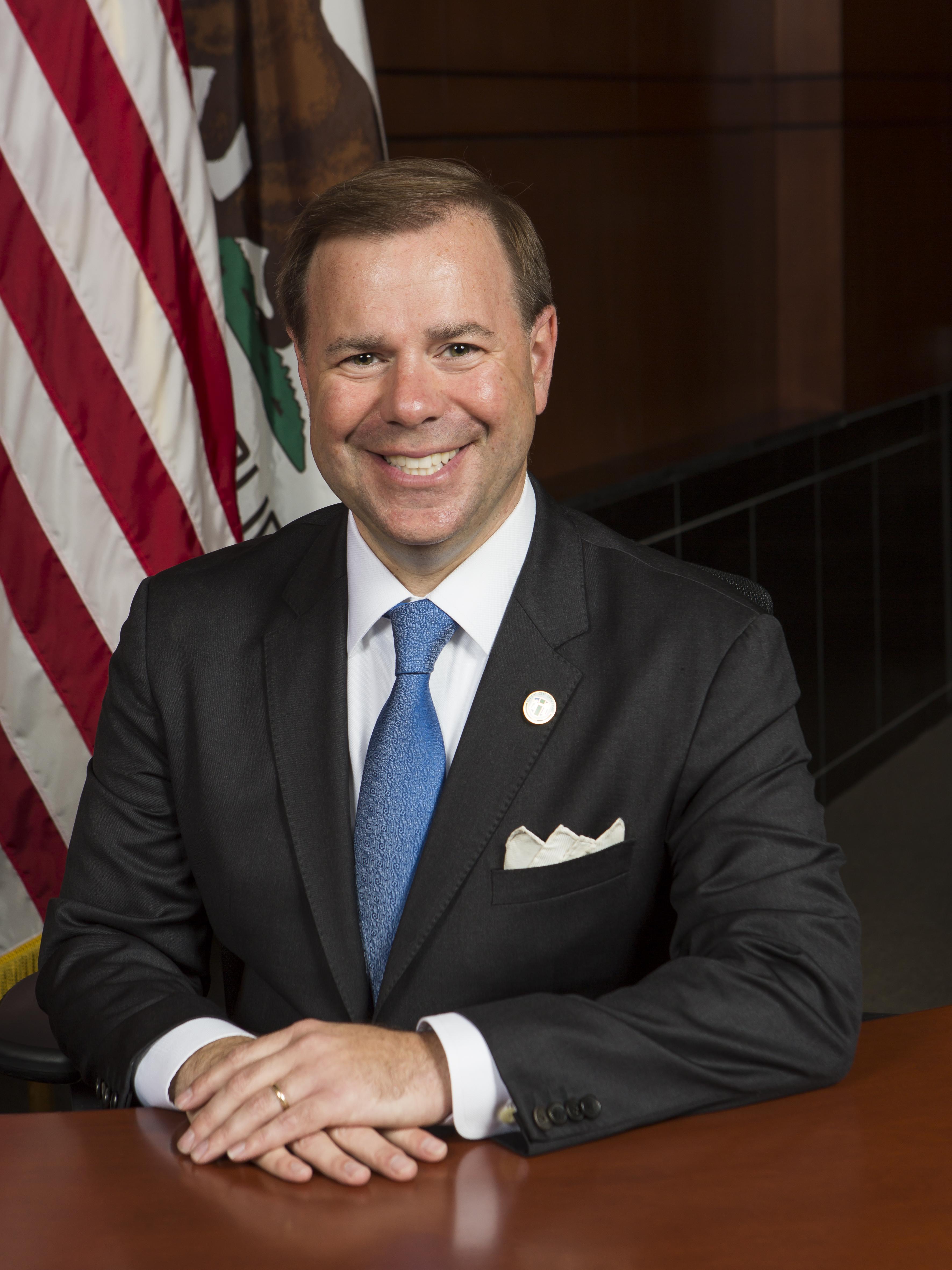 Smiling man in a dark suit and blue tie, seated at a desk, with an American flag behind him.