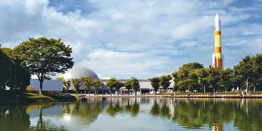 Park with a lake, a grey domed building, trees, and a tall yellow and orange rocket under a blue sky.
