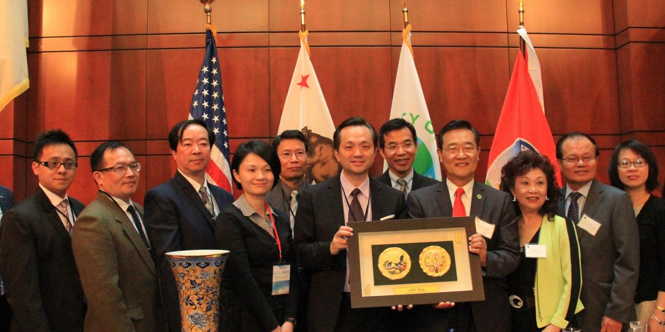 Group of adults in a formal room with flags behind them, holding a plaque