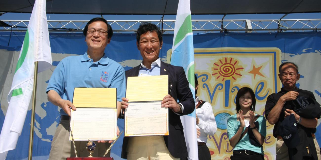 Two smiling men hold certificates on a stage with a blue backdrop and flags.