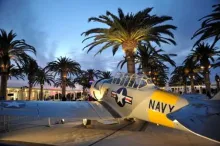 Vintage US Navy plane, yellow and gray, under palm trees at dusk.