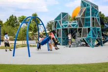 Two girls swing on a large blue swing in a modern park with geometric climbing structures.