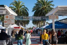 A festive outdoor Holiday Faire with crowds, stalls, a banner, and palm trees.