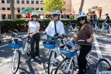 Three women wearing helmets and sunglasses pose with white and blue rental bikes.
