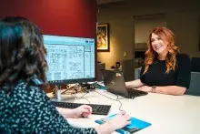 Two women are talking at a desk with a computer.