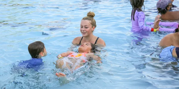 Young woman smiles, supporting a child floating on their back in a swimming pool.