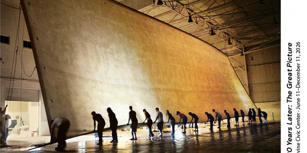 Photograph of workers collaboratively unrolling an enormous beige fabric in a vast hangar.
