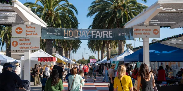 A festive outdoor Holiday Faire with crowds, stalls, a banner, and palm trees.