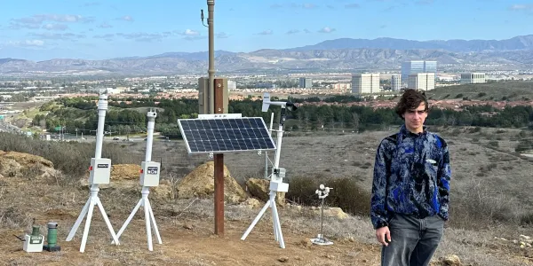 Person stands near scientific instruments and solar panel on a dry hill overlooking a city.