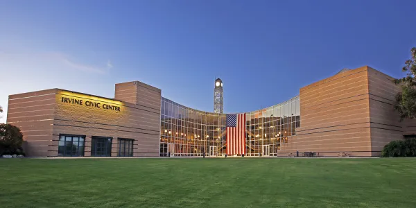 Modern Irvine Civic Center with glass facade, clock tower, and American flag over green lawn.