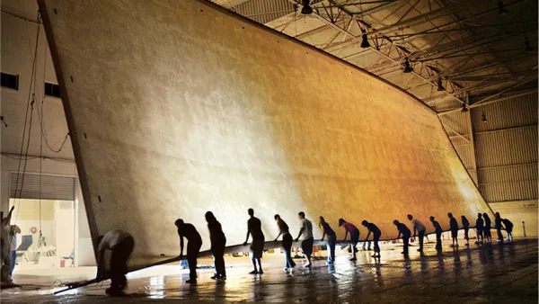 Photograph of workers collaboratively unrolling an enormous beige fabric in a vast hangar.
