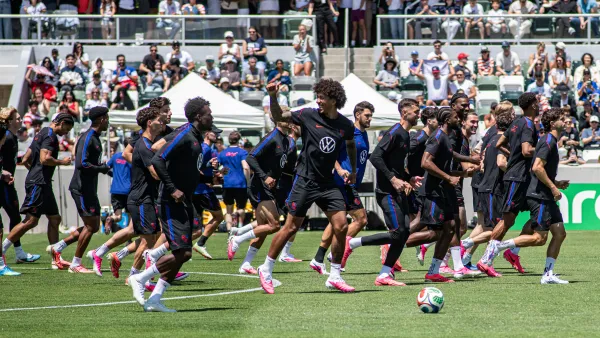 Soccer players in dark uniforms run across a green field with a stadium crowd behind them.