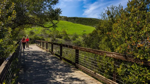 Two people walk on a bridge at Bommer Canyon. The bridge is surrounded by green trees and grassy hills are in the background.