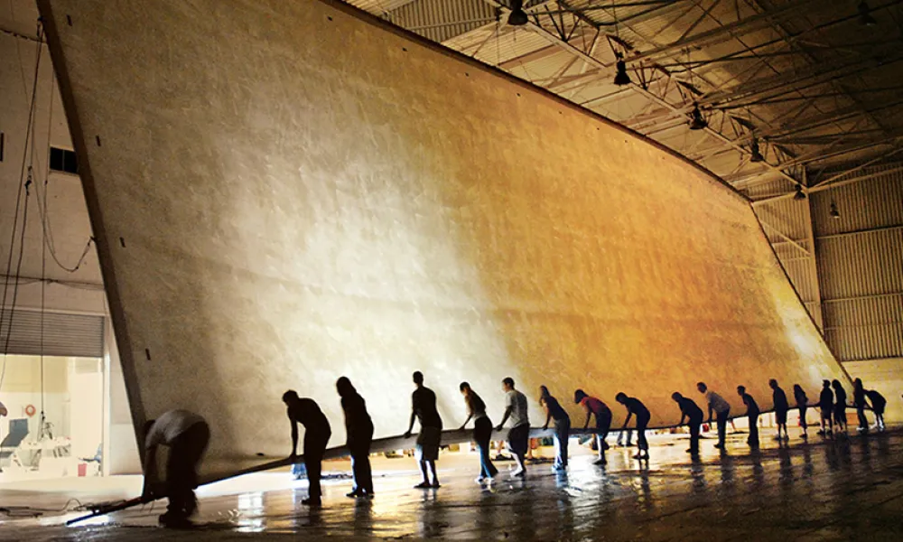 Photograph of workers collaboratively unrolling an enormous beige fabric in a vast hangar.