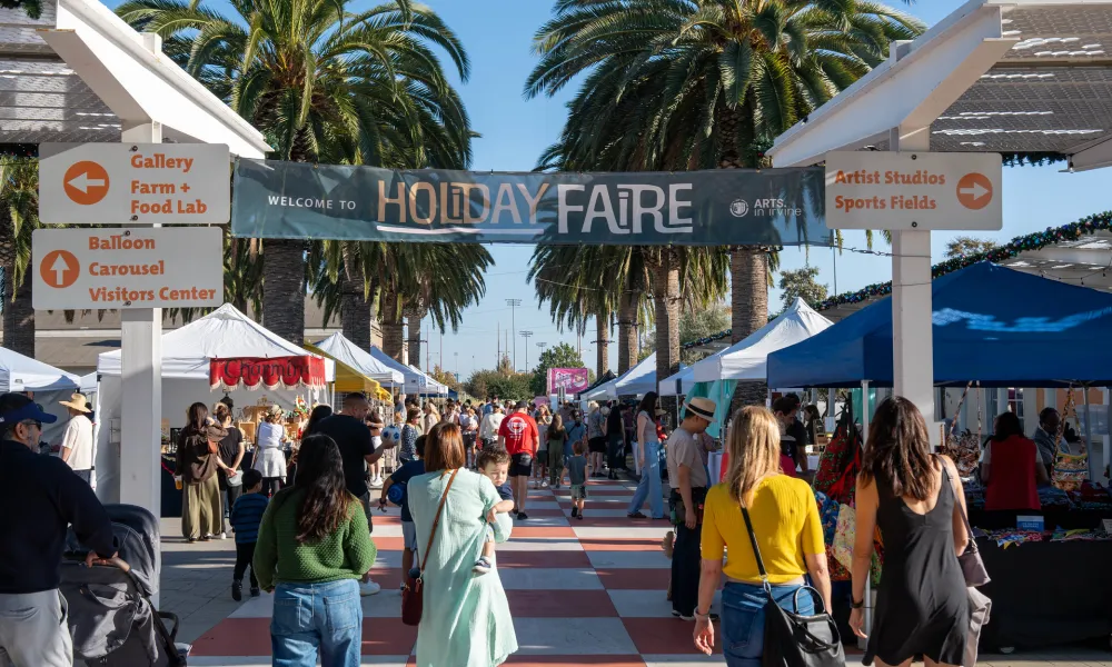 A festive outdoor Holiday Faire with crowds, stalls, a banner, and palm trees.