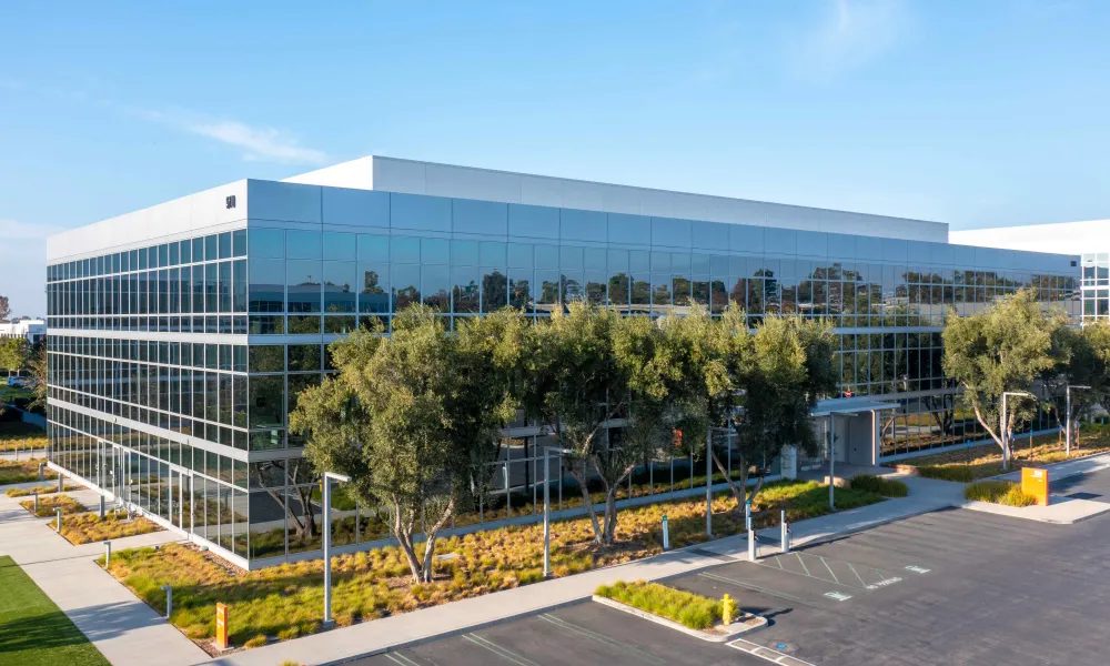 Modern glass office building with trees and landscaping under a blue sky.