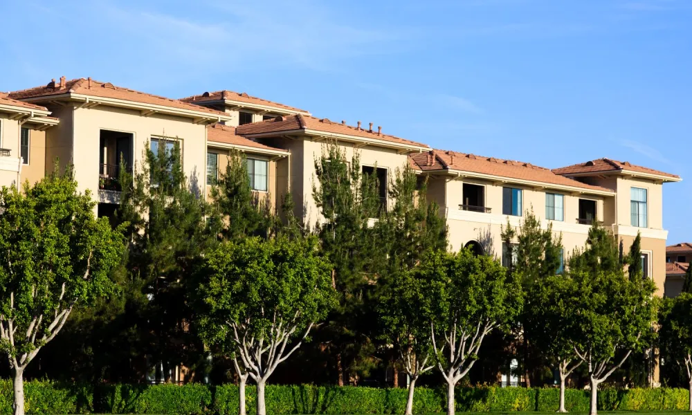 Tan apartment building with red tile roof, lush green trees, and blue sky.