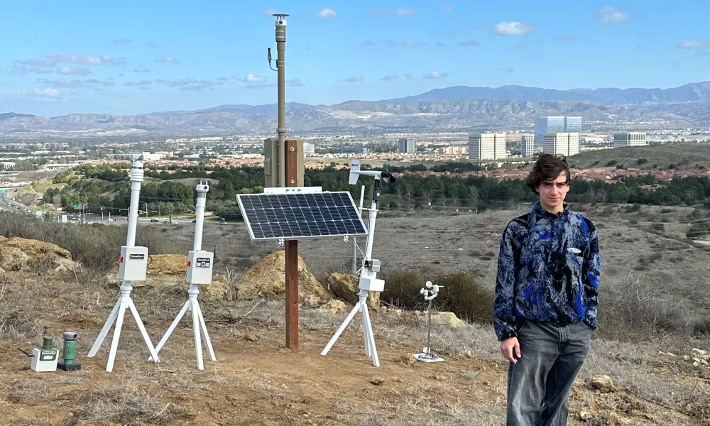 Person stands near scientific instruments and solar panel on a dry hill overlooking a city.