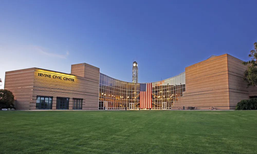 Modern Irvine Civic Center with glass facade, clock tower, and American flag over green lawn.