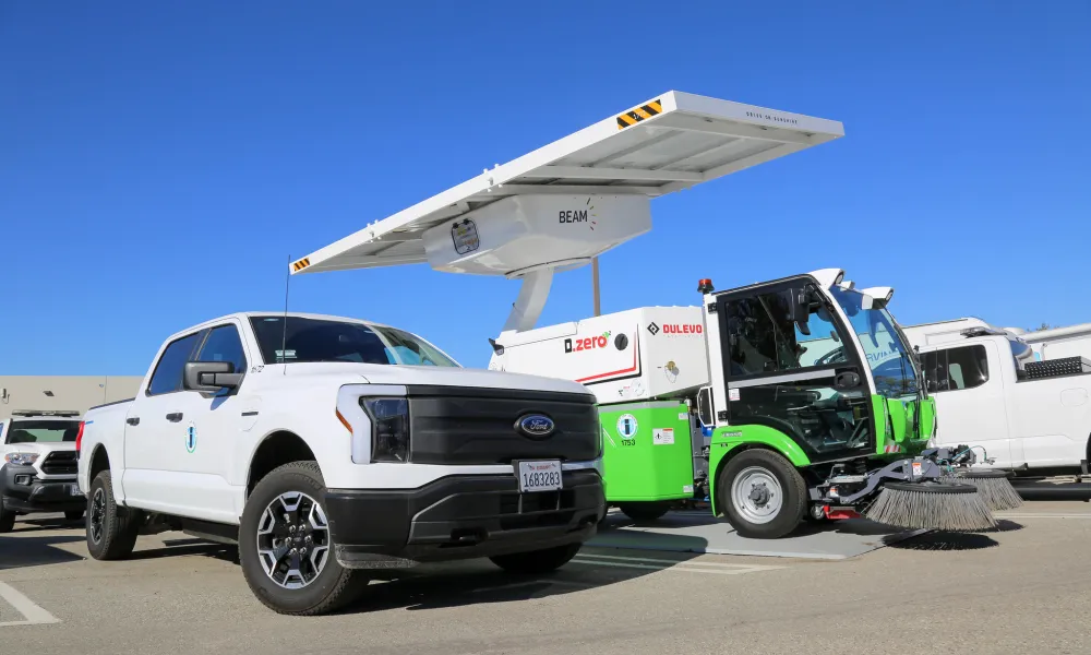 A white electric truck and green and white street sweeper are parked under a solar-powered EV charger.