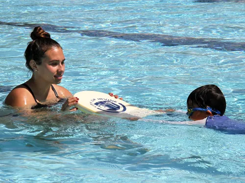 Woman teaching a child to swim with a kickboard in a bright blue pool.
