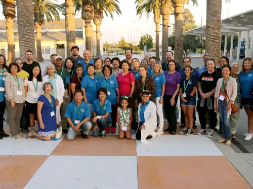 Large, diverse group of people stand outdoors among palm trees and modern buildings.