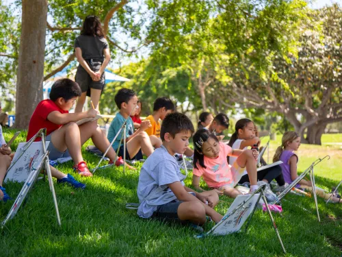 Young children sit on a grassy hill, drawing on small easels outdoors with an instructor.