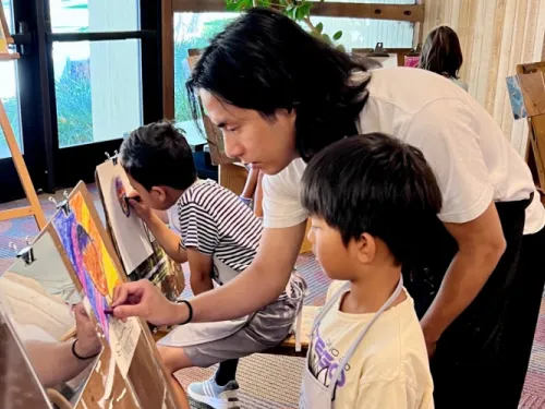 Adult instructor guides a young boy painting a colorful artwork in an art class.