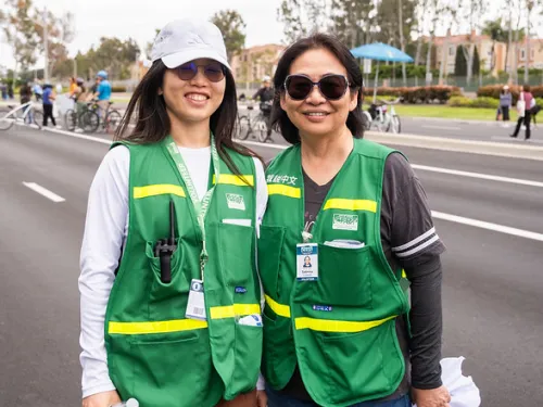 Two smiling women in green safety vests stand on a road; people and trees in background.