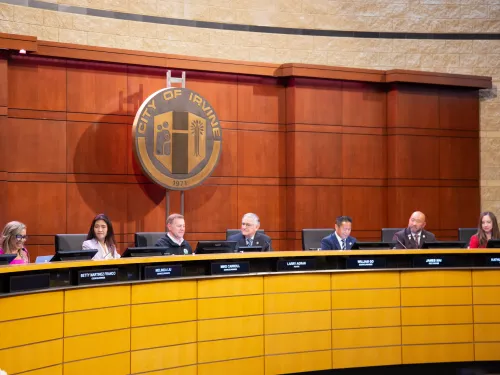 Panel of seven people seated at a long dais in a council chamber under a large city seal.