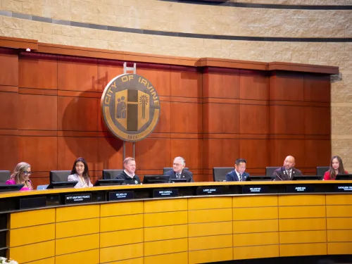 Panel of seven people seated at a long dais in a council chamber under a large city seal.