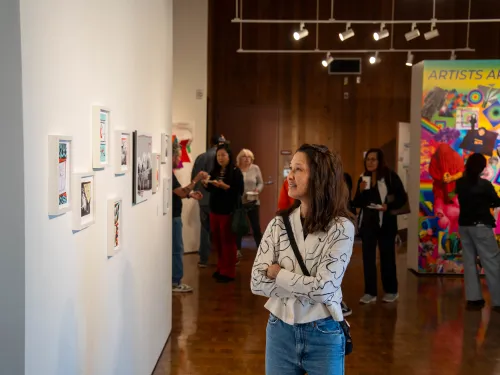 Woman smiles, looking at framed art in a gallery; colorful exhibit in background.