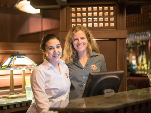 Two smiling women, restaurant staff, stand behind a wooden counter.