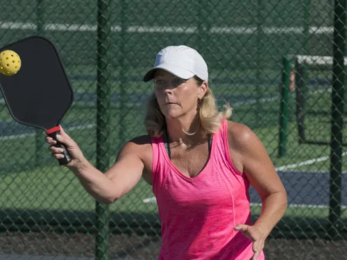 Woman with a white cap and pink tank top holding a racquet and about to hit a pickeball