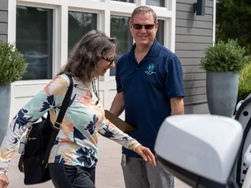 Smiling man in blue polo assists woman in colorful shirt getting into a white car at a shopping center