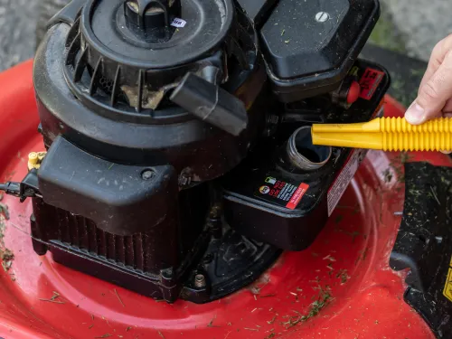 Hand pouring oil from a yellow funnel into a red lawnmower engine.