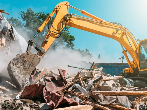 Yellow excavator demolishes a building, sending dust and debris into the air.