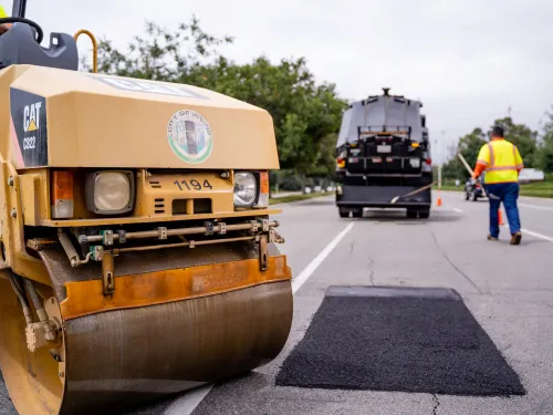Yellow road roller poised over fresh asphalt patch, road worker and truck nearby.