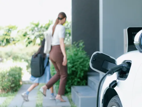 White electric car charging outside a house; woman and child walk inside.