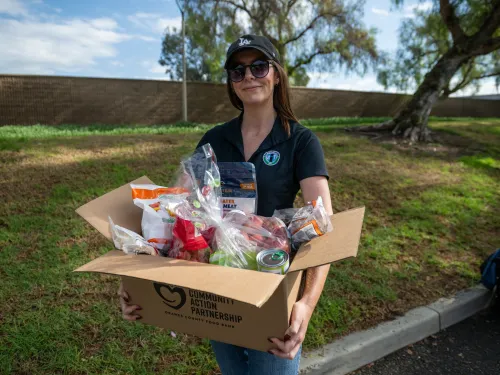 Smiling woman in a cap holds a full box of groceries outdoors.