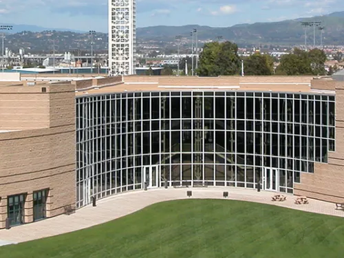 Modern tan building with large glass entrance, tall spire, and a pyramidal roof. Green lawn foreground, distant hills.
