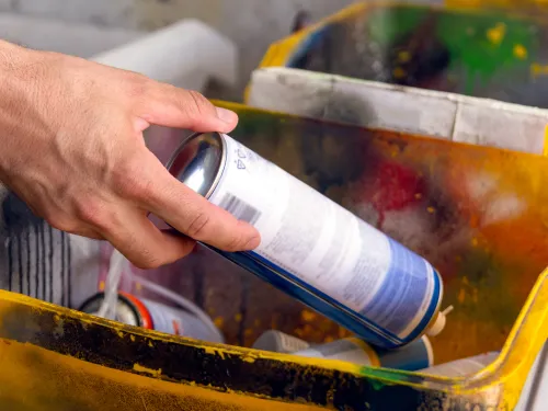 Hand places a blue spray paint can into a yellow bin covered in colorful paint splatters.