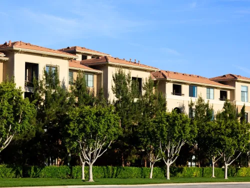 Multi-story tan building with terracotta roofs, green trees, and blue sky.