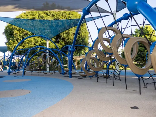 Modern playground with blue climbing structures, tan rings, soft ground, and shade sails.